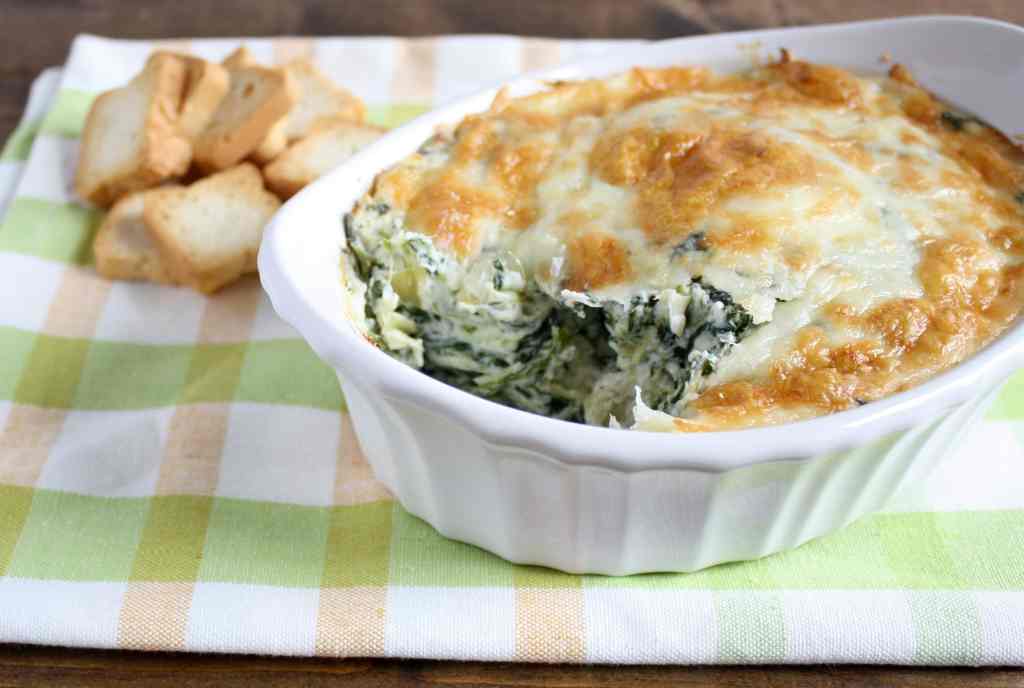 No mayo spinach artichoke dip in a white baking dish on top of a checkered tea towel. Crackers rest in the background. 