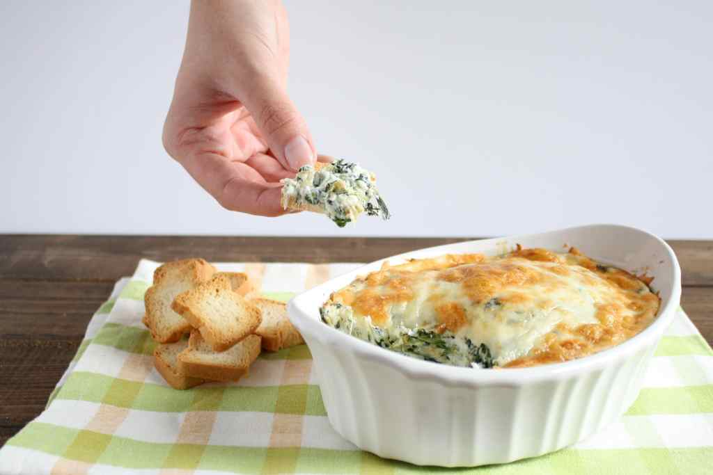 No mayo spinach artichoke dip being scooped out of a white baking dish that rests on a tea towel. 