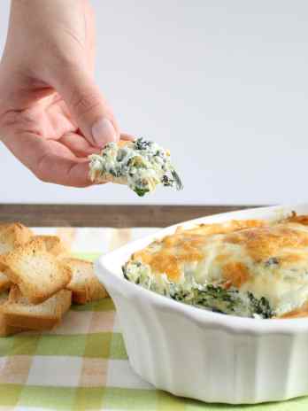 No mayo spinach artichoke dip being scooped out of a white baking dish that rests on a tea towel.