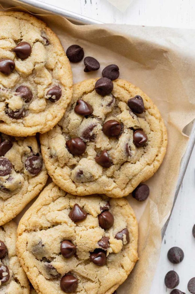 A close up image of chocolate chip cookies on a baking sheet lined with brown parchment paper.