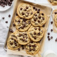 A pile of chocolate chip cookies on a baking sheet lined with parchment paper surrounded by more cookies and chocolate chips.