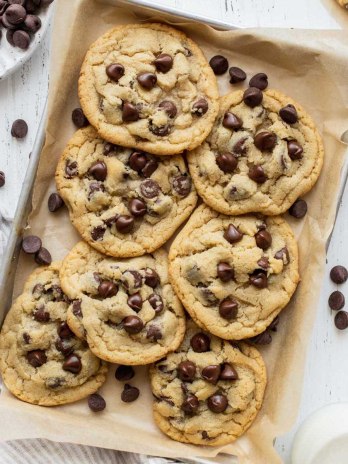 A pile of chocolate chip cookies on a baking sheet lined with parchment paper surrounded by more cookies and chocolate chips.