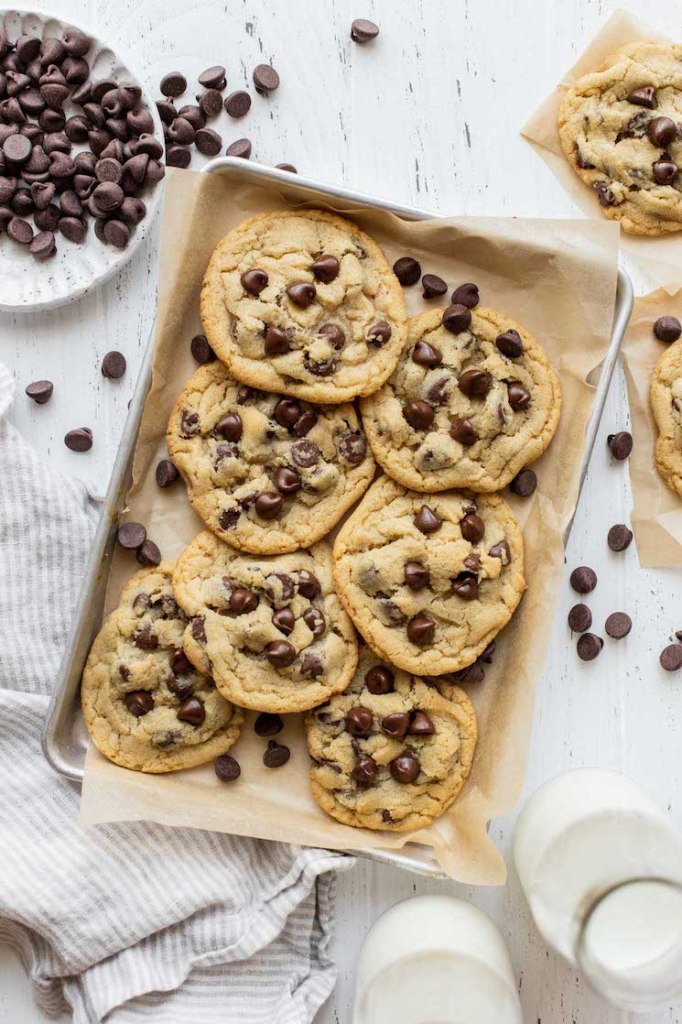 A pile of chocolate chip cookies on a baking sheet lined with parchment paper surrounded by more cookies and chocolate chips.