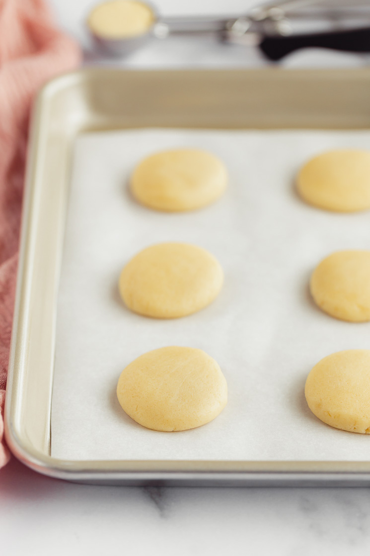 Scoops of sugar cookie dough, on a metal baking pan lined with parchment paper, flattened and ready to go into the oven.