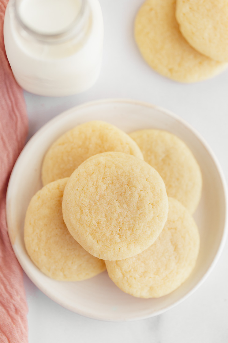 A white plate with sugar cookies and more cookies and a jug of milk in the background.