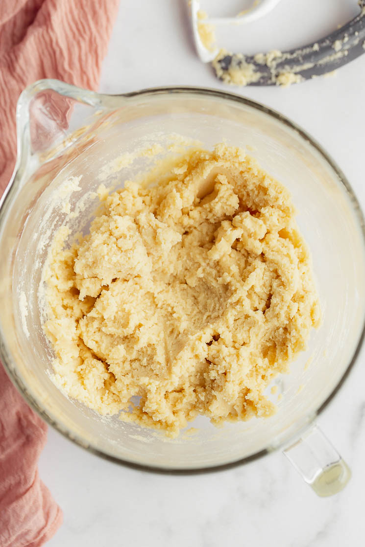 A glass mixing bowl with sugar cookie dough with a beater and a pink napkin in the background.