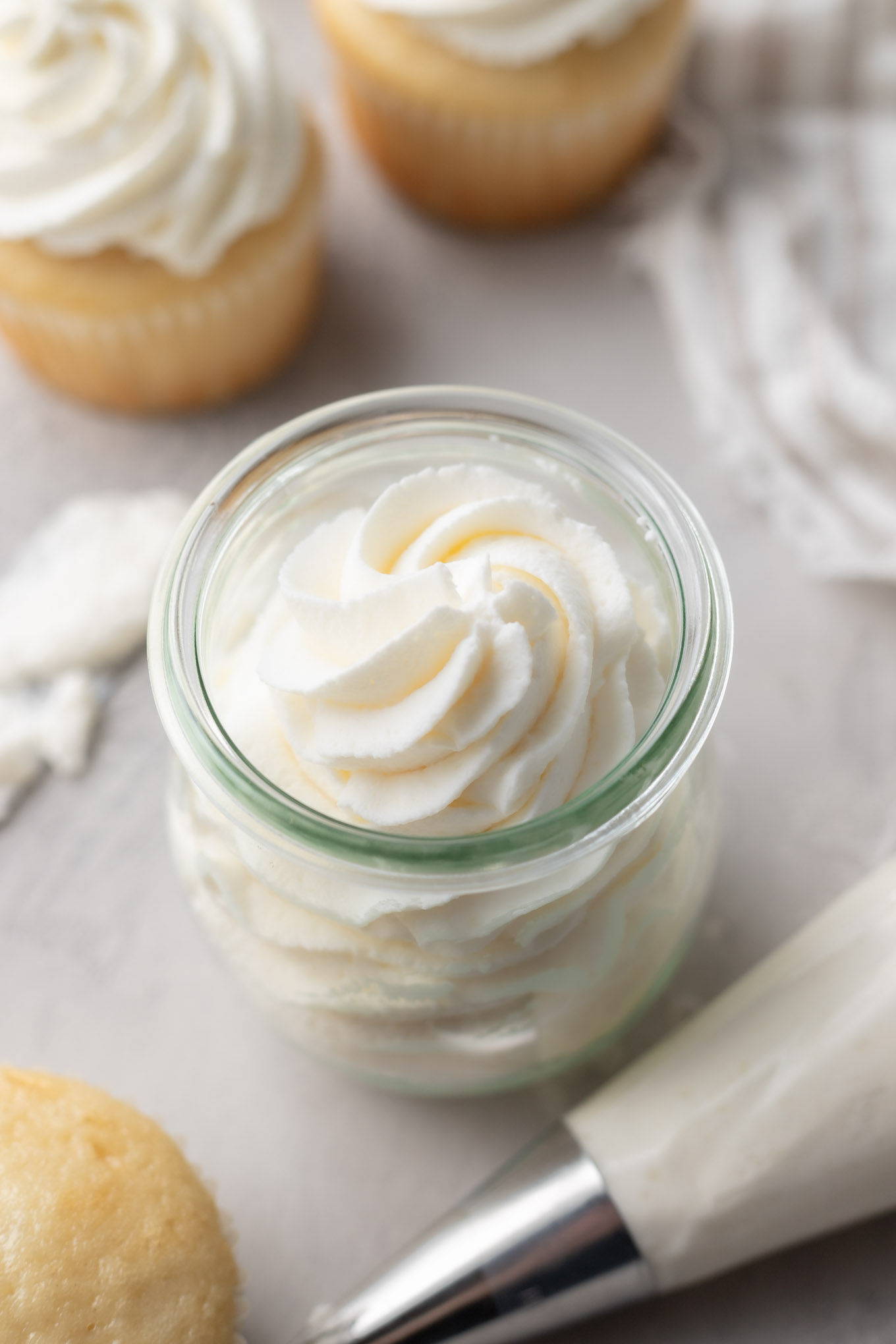 An overhead view of stabilized whipped cream in a glass jar, next to vanilla cupcakes. 