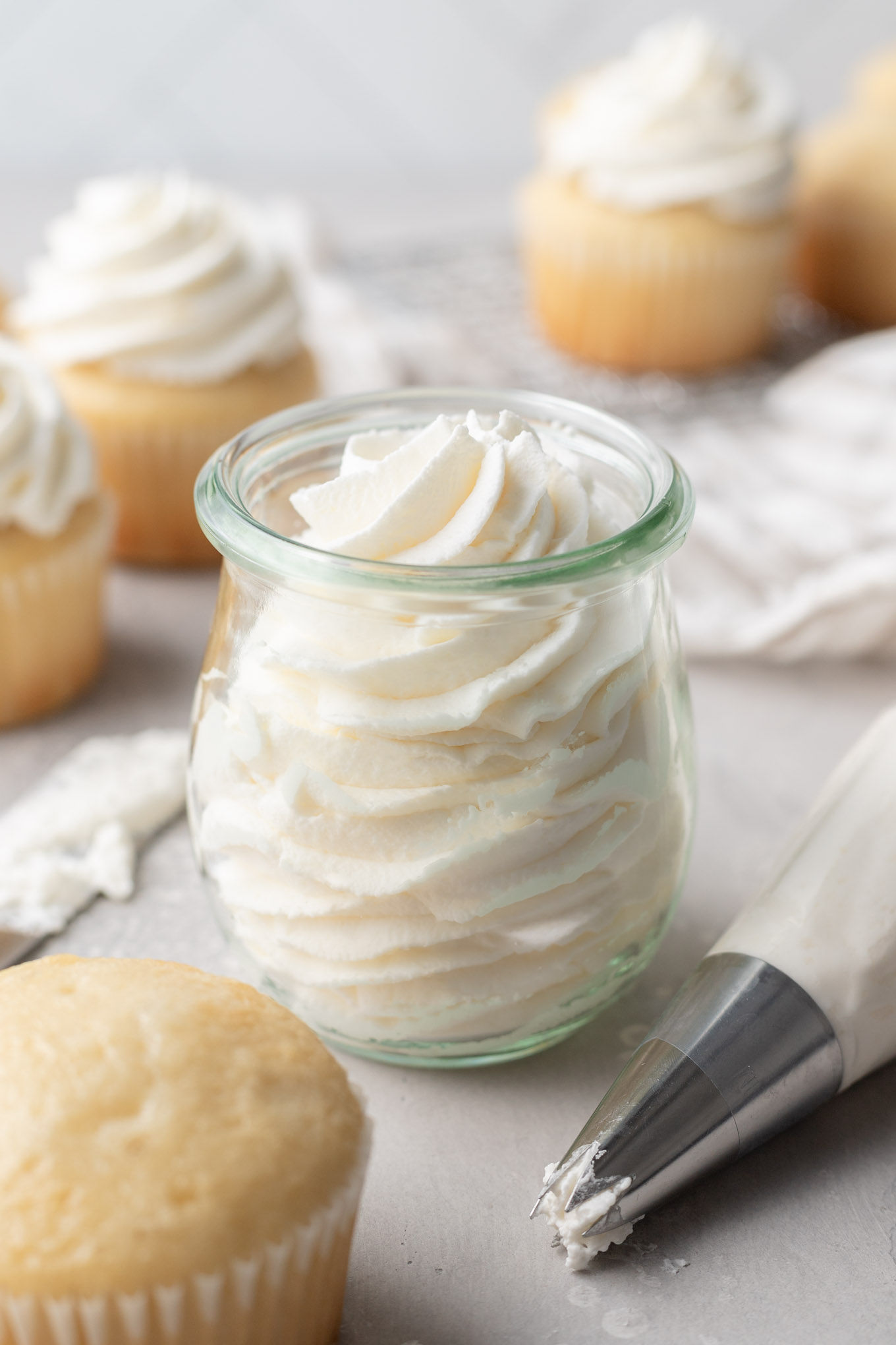 A glass jar filled with whipped cream. Vanilla cupcakes surround the jar. 