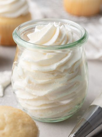 A glass jar filled with gelatin whipping cream. Vanilla cupcakes surround the jar.