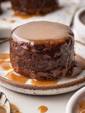 A small round date cake covered in toffee sauce on a white speckled plate.