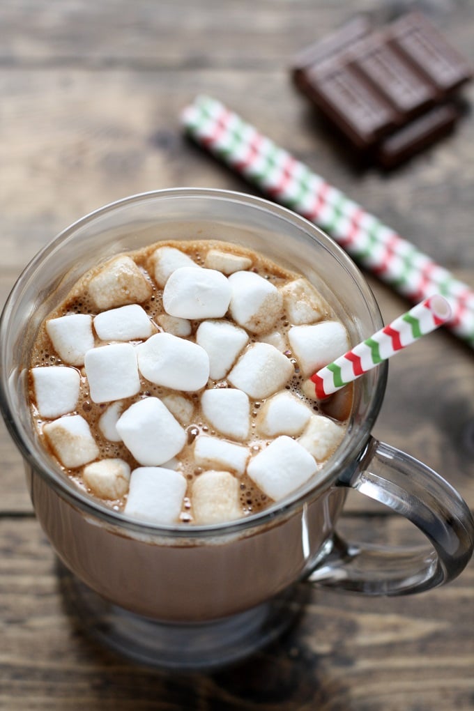 Mug of homemade hot chocolate topped with mini marshmallows and a straw. Chocolate squares rest in the background. 