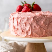 A strawberry cake with strawberry buttercream frosting sitting on top of a marble cake stand. Several fresh strawberries rest on top of the cake.