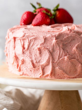 A strawberry cake with strawberry buttercream frosting sitting on top of a marble cake stand. Several fresh strawberries rest on top of the cake.