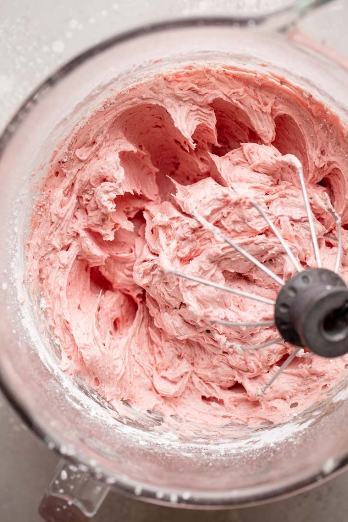 A glass mixing bowl filled with strawberry buttercream frosting.