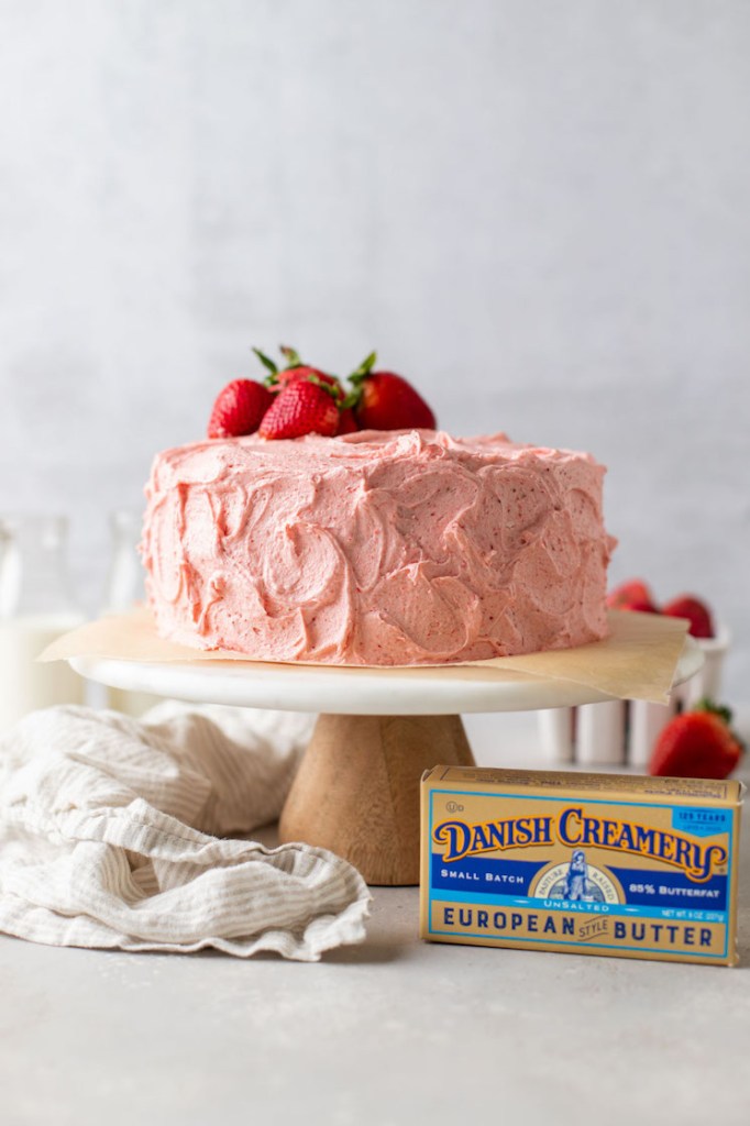 A strawberry cake topped with fresh strawberries sitting on top of a cake stand. A box of butter and a napkin are sitting next to the cake stand.