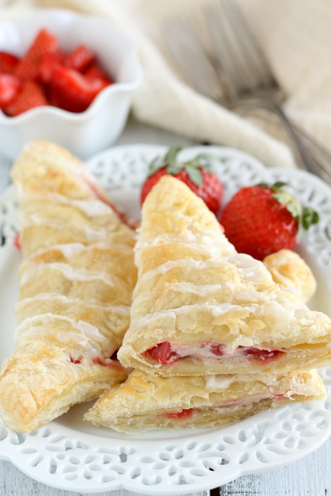 Two strawberry cream cheese turnovers on a white plate. One turnover has been cut in half and stacked on the other half to show the inside of it. Two strawberries rest on the plate and some chopped strawberries and forks rest in the background.