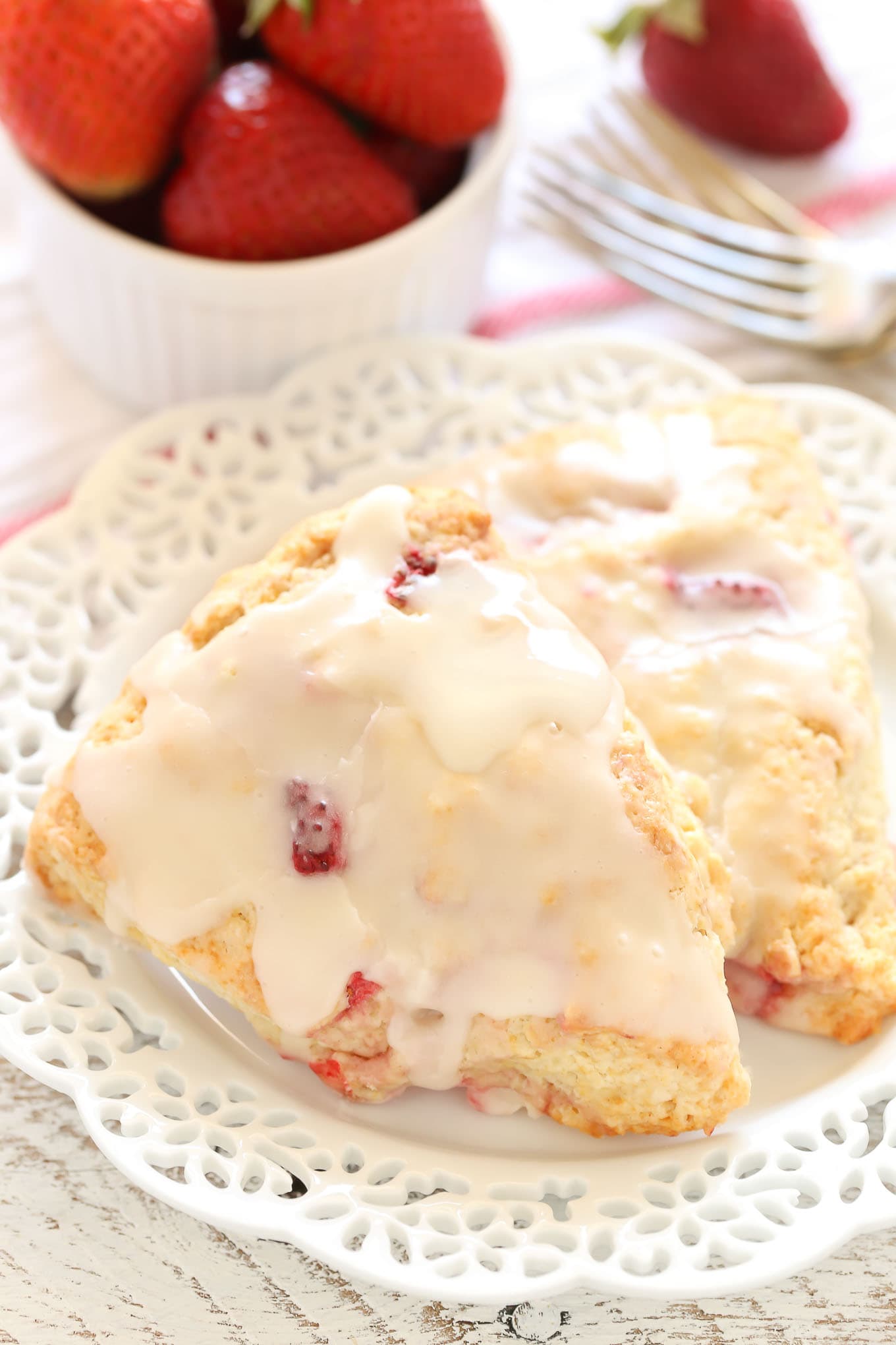 Two glazed strawberry scones on a white plate. A small bowl of berries and two forks rest in the background. 