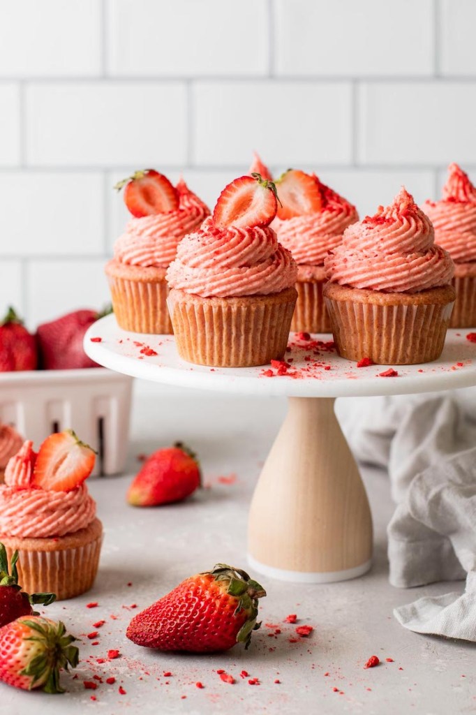 A side view of fresh strawberry cupcakes on a cake stand. Additional cupcakes and fresh berries litter the counter underneath. 