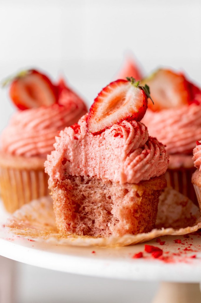 A close up view of cupcakes with strawberry frosting on a cake stand. The front cupcake has a bite missing. 