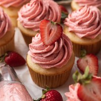 Several vanilla cupcakes topped with strawberry buttercream frosting. A piping bag sits next to the cupcakes.