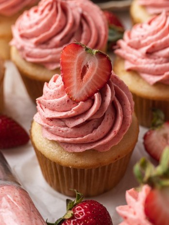 Several vanilla cupcakes topped with strawberry buttercream frosting. A piping bag sits next to the cupcakes.