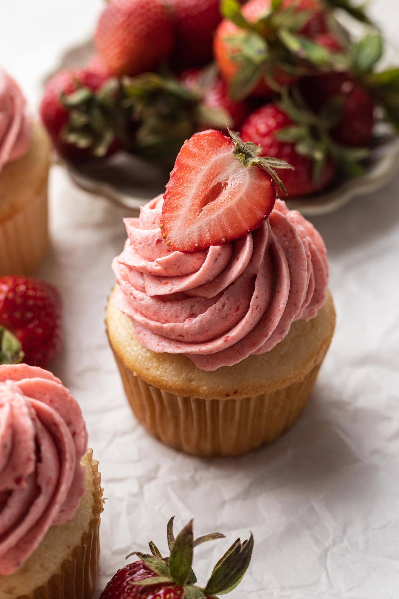 A vanilla cupcake topped with strawberry frosting and garnished with a fresh strawberry half. 