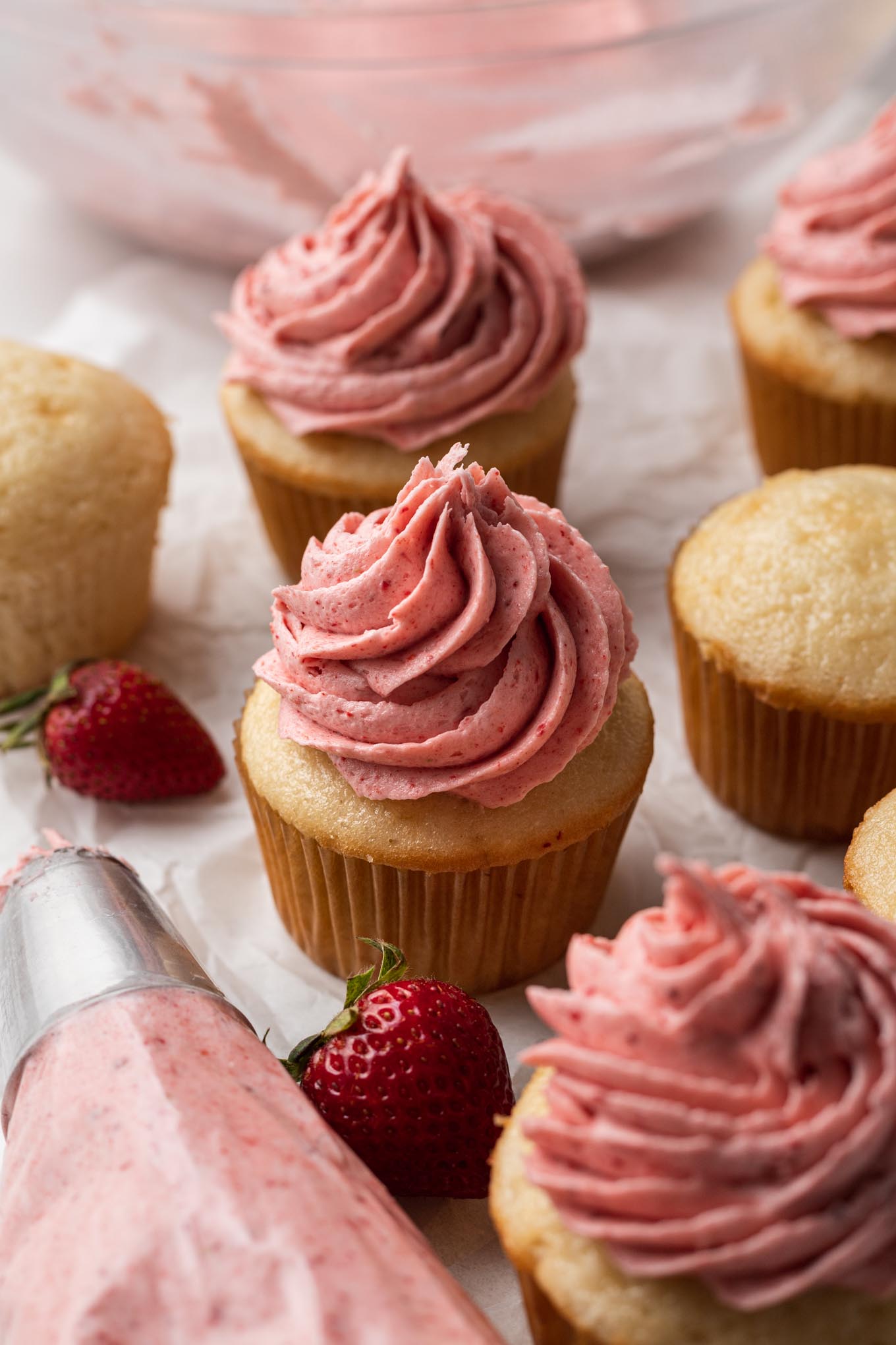 A cluster of vanilla cupcakes, some with frosting and some plain. A piping bag of frosting rests in the foreground. 