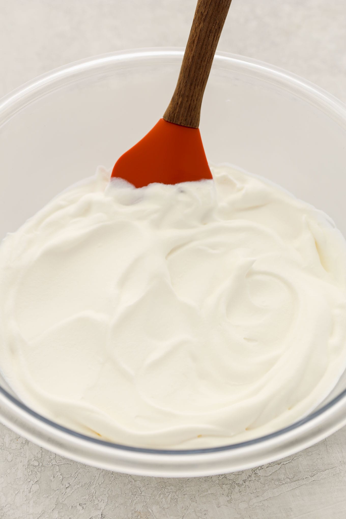 A glass bowl filled with homemade whipped cream and a rubber spatula in it.