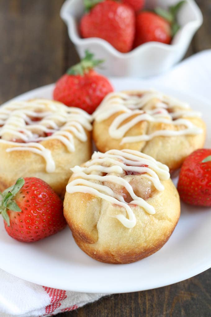 Three strawberry rolls topped with cream cheese frosting on a white plate with fresh berries. 