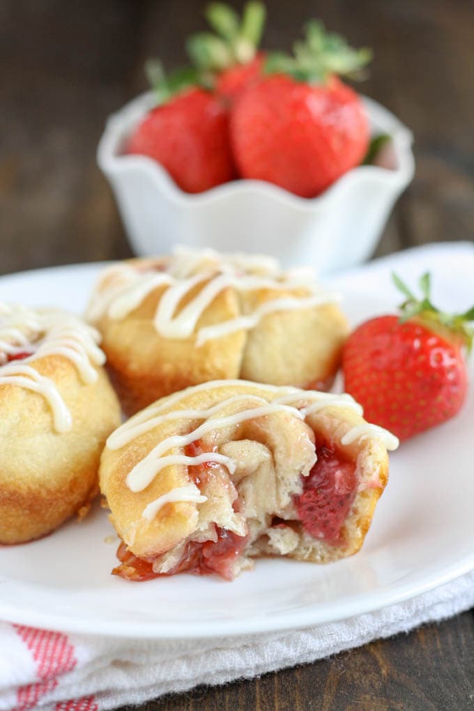 Three strawberry cinnamon rolls on a white plate. The front sweet roll has a bite missing. There's a bowl of fresh berries in the background. 