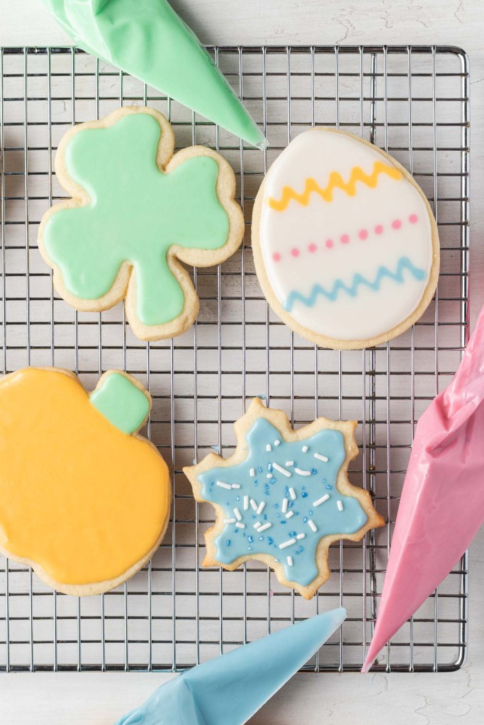 An overhead view of four iced sugar cookies on a wire cooling rack. Three piping bags filled with icing surround the cookies. 