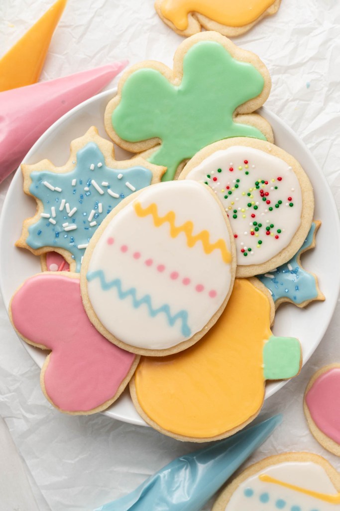 An overhead view of a pile of iced sugar cookies on a white plate. Piping bags of colored sugar cookie frosting are strewn around the plate. 