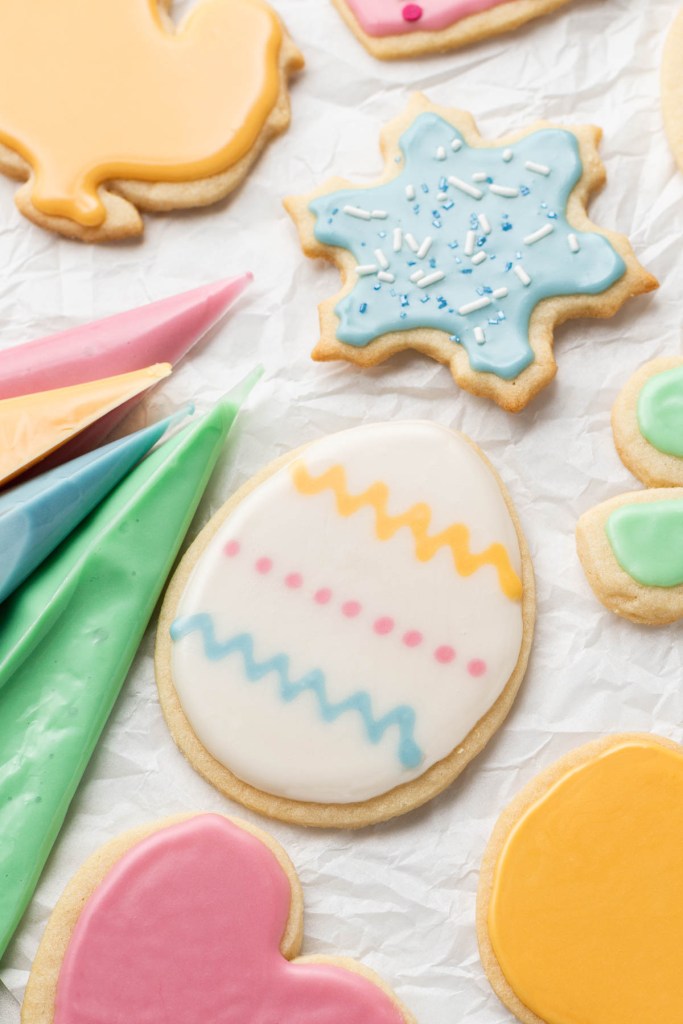 Piping bags of colored icing and iced sugar cookies on white parchment paper. 