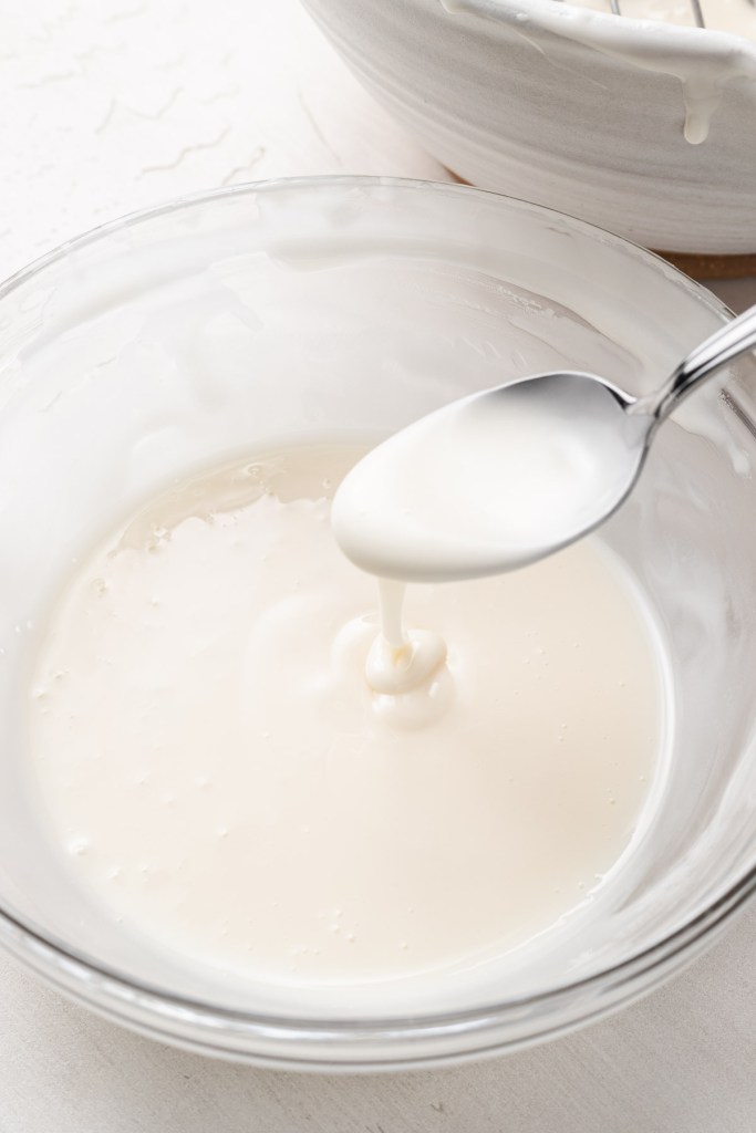 A glass mixing bowl of flood icing with a spoon held aloft. 