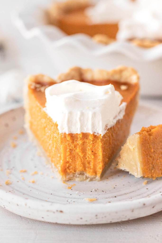 A slice of sweet potato pie topped with whipped cream on a white speckled dessert plate. A bite is missing. The rest of the pie rests in the background. 