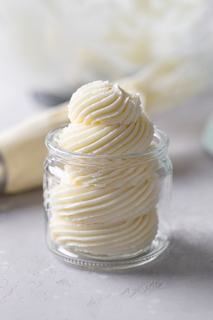 Swiss meringue buttercream frosting piped into a glass jar. A piping bag lies in the background. 