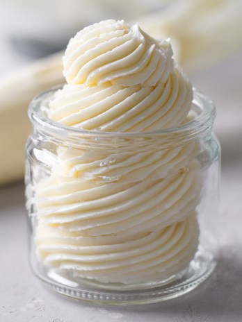 Swiss meringue buttercream frosting piped into a glass jar. A piping bag lies in the background.