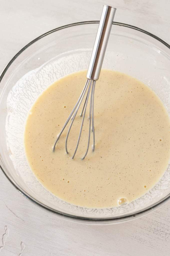 Buttermilk, sour cream, eggs, and vanilla mixed together in a glass mixing bowl with a metal whisk.