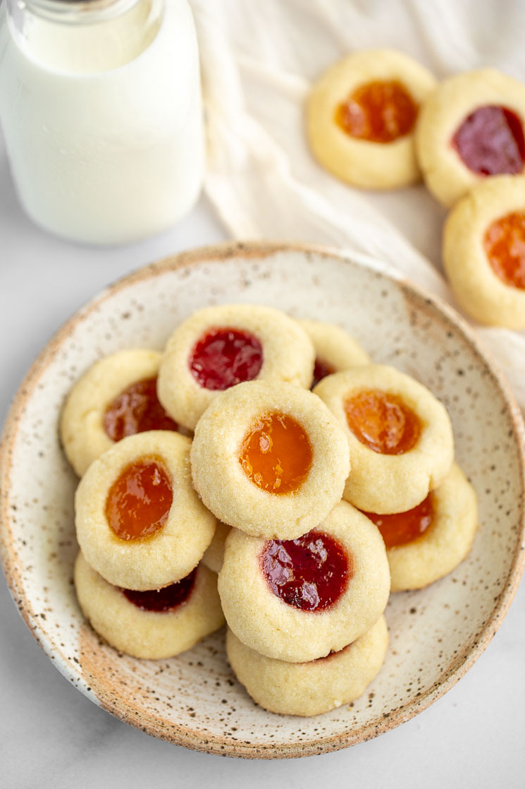An speckled plate filled with thumbprint cookies with more cookies and milk behind it.
