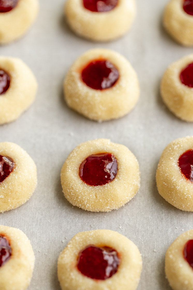 A batch of unbaked thumbprint cookies filled with jam lined up on a piece of parchment paper.