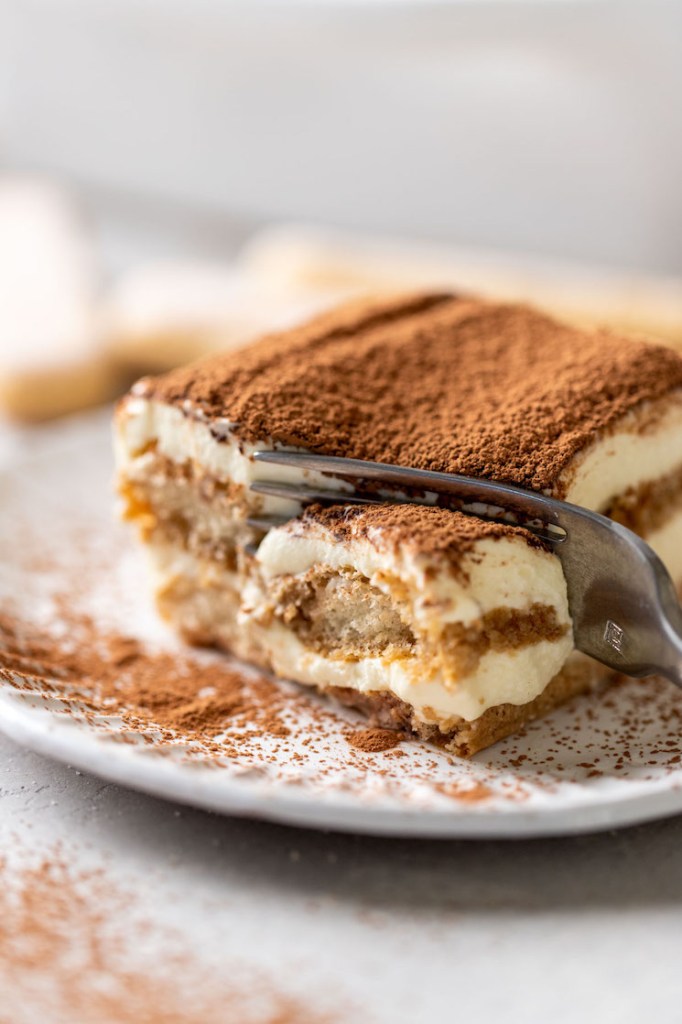 A slice of tiramisu on a white decorative plate. A fork is digging into the end of the slice.
