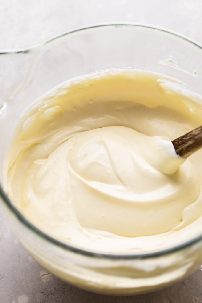 A glass mixing bowl with the finished mascarpone filling after the heavy cream has been folded in. A rubber spatula with a wooden handle rests in the bowl.