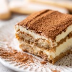 A slice of tiramisu on a white decorative plate. Three lady fingers rest in the background.
