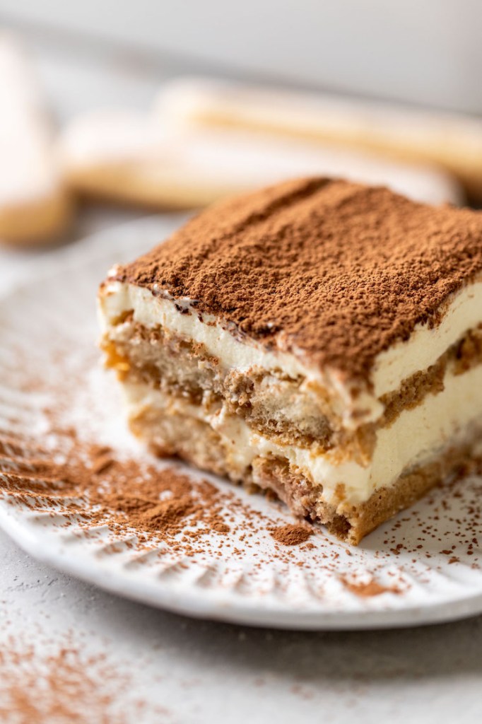 A slice of tiramisu on a white decorative plate. Three lady fingers rest in the background.