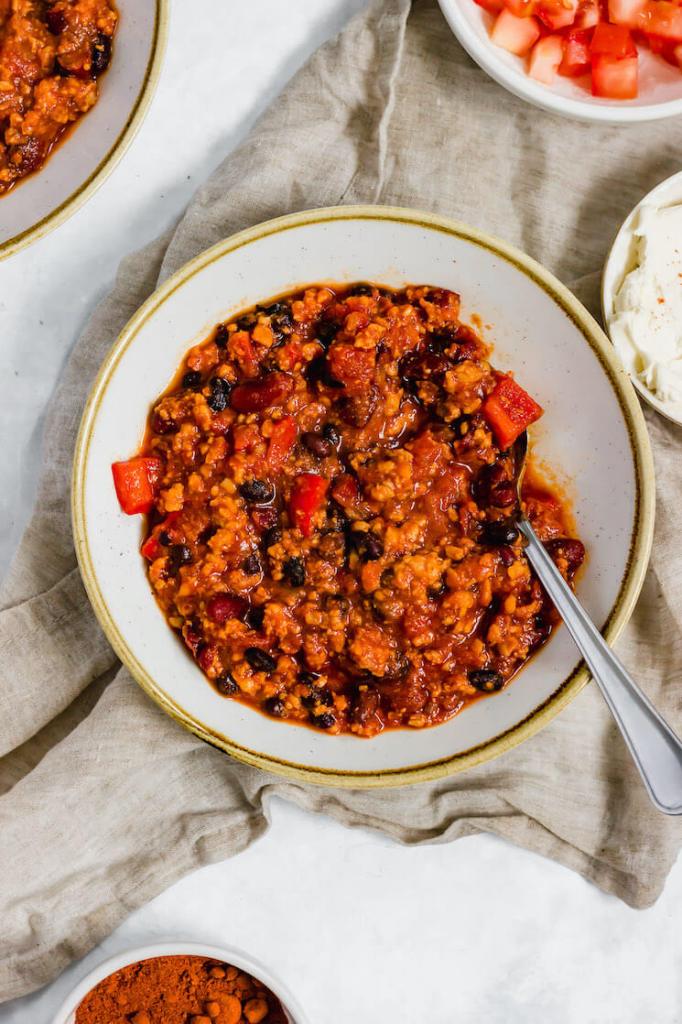 A bowl of turkey chili with a spoon in it. Several bowls with sour cream, diced tomatoes, and more chili are around it.