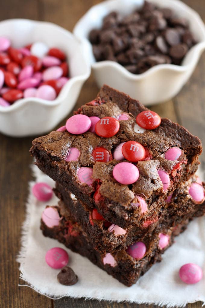 A stack of three Valentine brownies on a square of fabric. A bowl of M&M's and a bowl of chocolate chips are in the background.