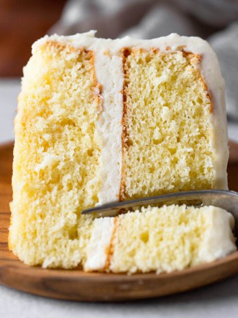 A slice of vanilla cake lying on its side on a wood plate. A fork is digging into the end of the slice.