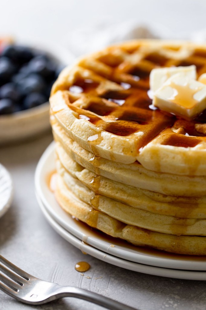 A side view of a stack of fluffy waffles topped with butter and syrup. A bowl of fresh berries rests in the background. 