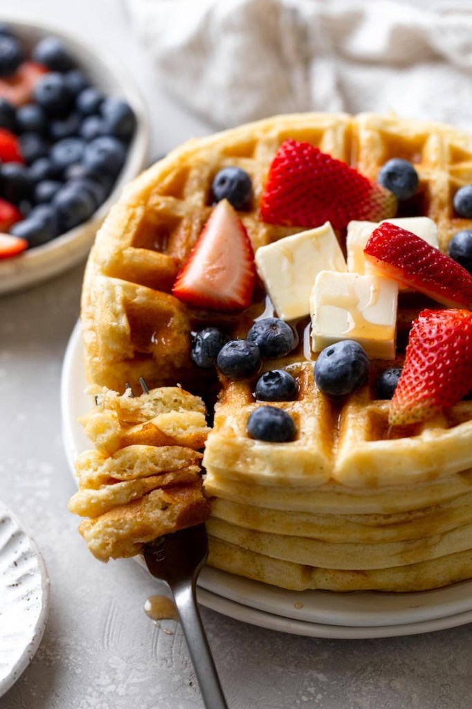 A tall stack of waffles topped with berries, butter, and syrup. A fork rests on the plate as well, speared with a big bite of waffle. 