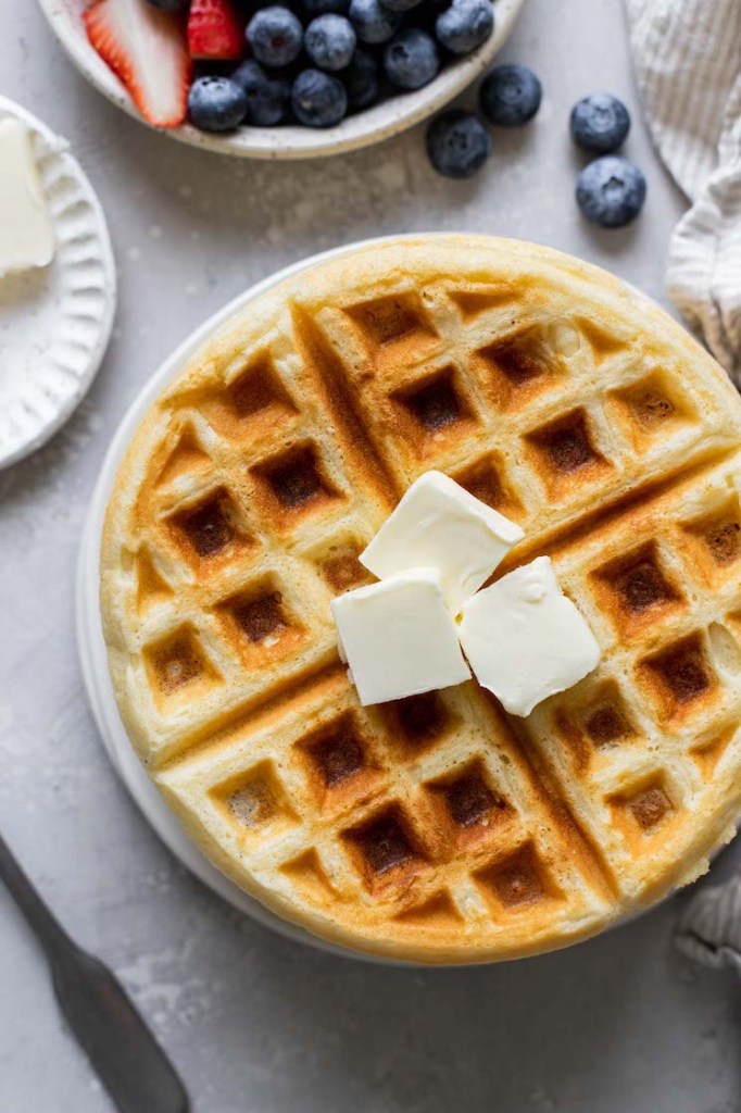 An overhead view of a waffle topped with three pats of butter. A dish of fresh berries rests on the side. 